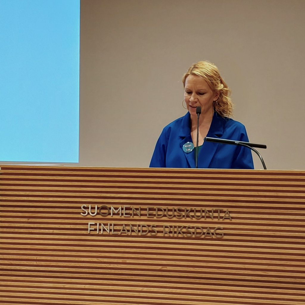Siniseen asuun pukeutunut Kaari Mattila seisoo puhujakorokkeen takana. Korokkeessa on teksti Suomen eduskunta Finlands riksdag. 